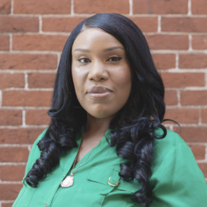 A headshot of Cattima in front of a brick wall. She is wearing a green shirt.