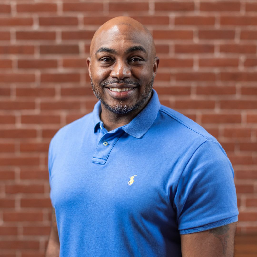 isaiah price in a blue shirt smiling against a brick wall